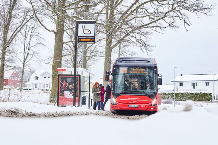 Bilden visar en röd stadsbuss som plockar upp resenärer vid en busshållplats. Marken är täckt av snö.