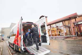 Bilden visar en förare hjälpa en resenär i rullstol upp för en ramp och in i en buss utanför entrén till Länssjukhuset Ryhov.