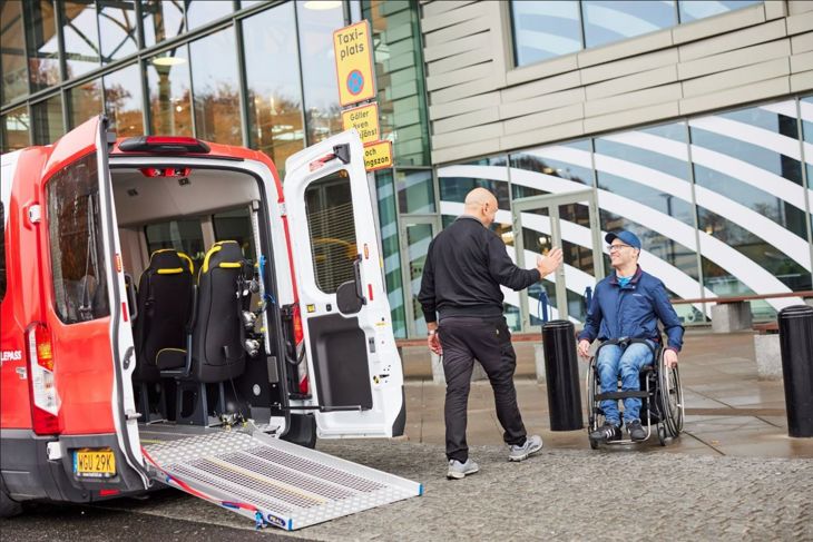 bild på färdtjänstbil och förare som hämtar upp kund i rullstol vid köpcentrum.