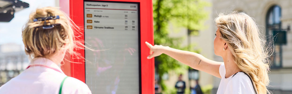 Två resenärer står och väntar på bussen och tittar på en hållplatsskylt.