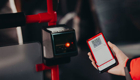 En hand håller i en mobiltelefon och på skärmen syns en biljett i appen. Biljetten hålls upp mot en validator ombord på en buss.