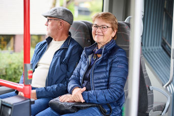 Två resenärer ombord på en buss. En man i keps som tittar ut genom fönstret och en kvinna som ler mot kameran.